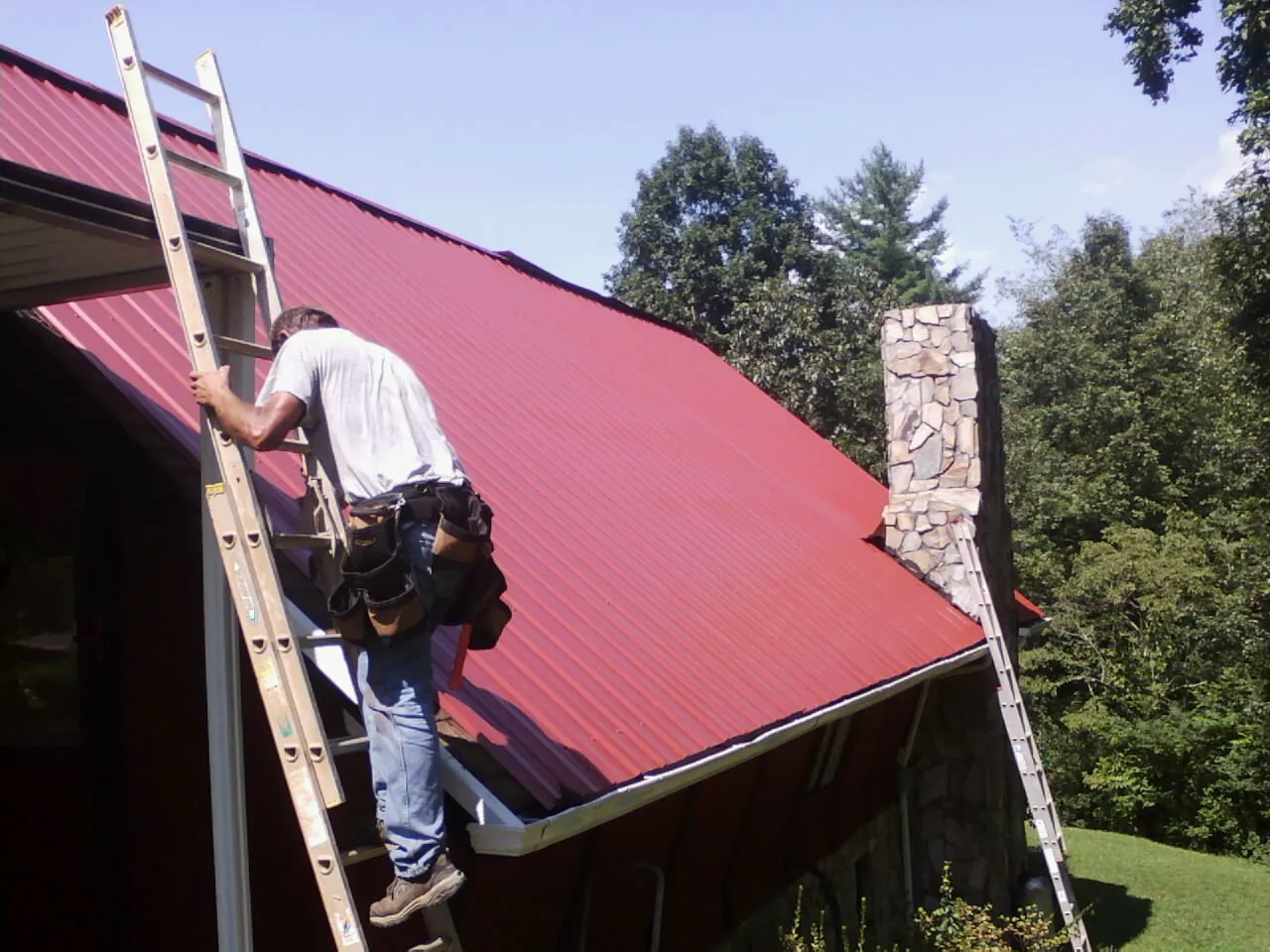 Roofer on a ladder beside a red metal roof and stone chimney — Soffit & Fascia Repair by Roofing Brothers in Bolivia